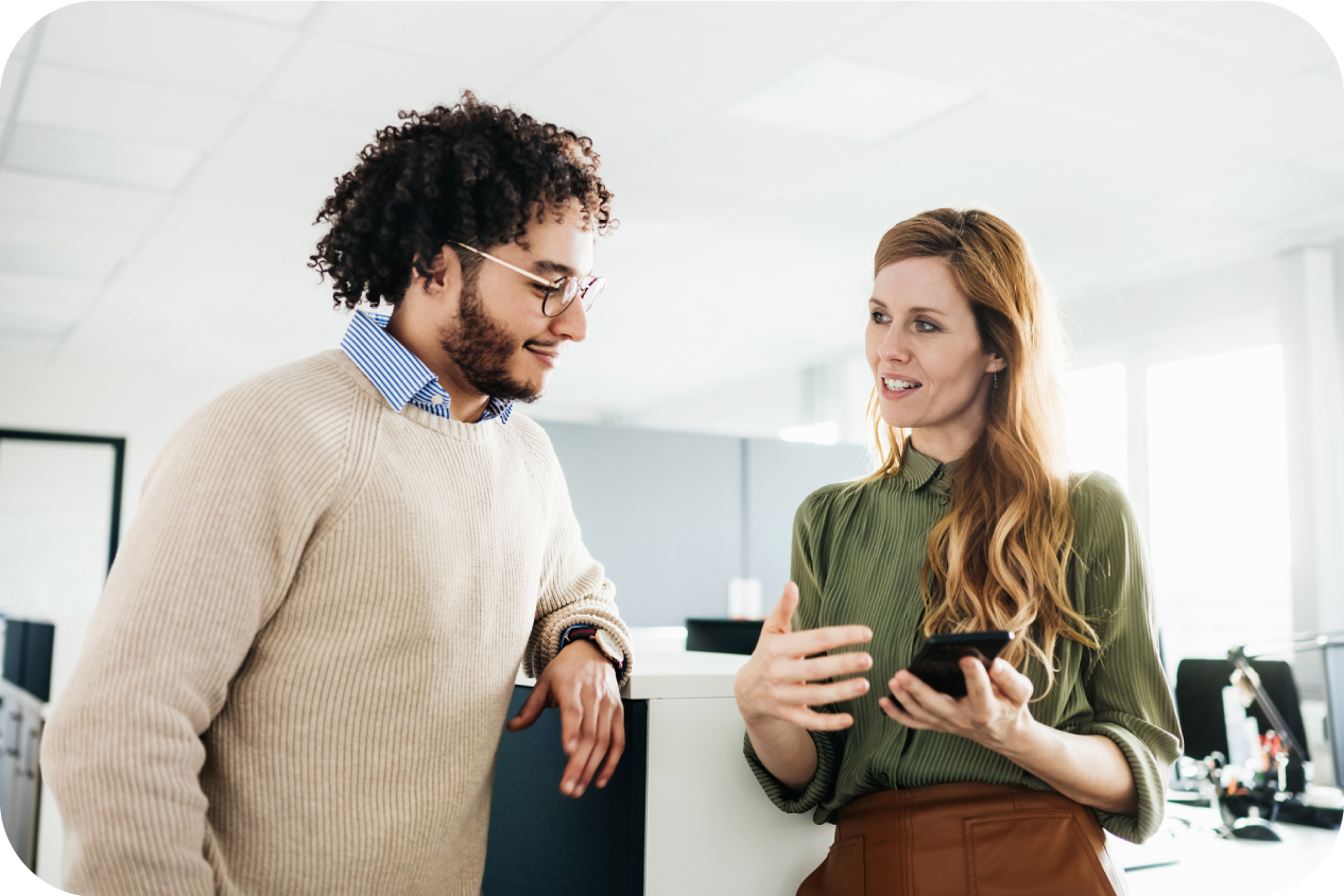 A woman is holding a smartphone and appears to be describing something to a male colleague in an office setting.