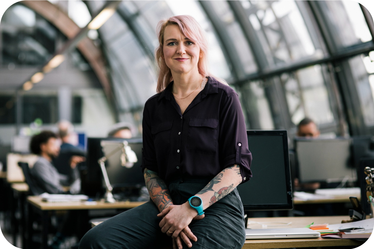 A smiling woman is sitting on her desk and looking into the camera. She has pink hair and various visible tattoos.