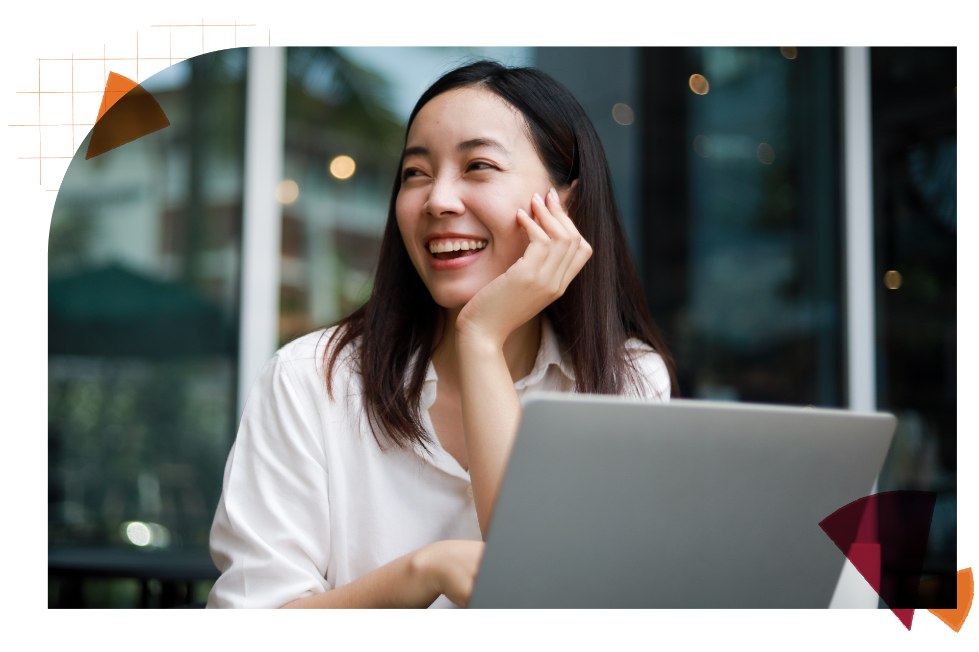 An asian woman with brunette mediem length hair is sitting outside with her laptop open and sitting at a table. She is smiling with her hand propped up against her chin and looking away from the camera towards the left.