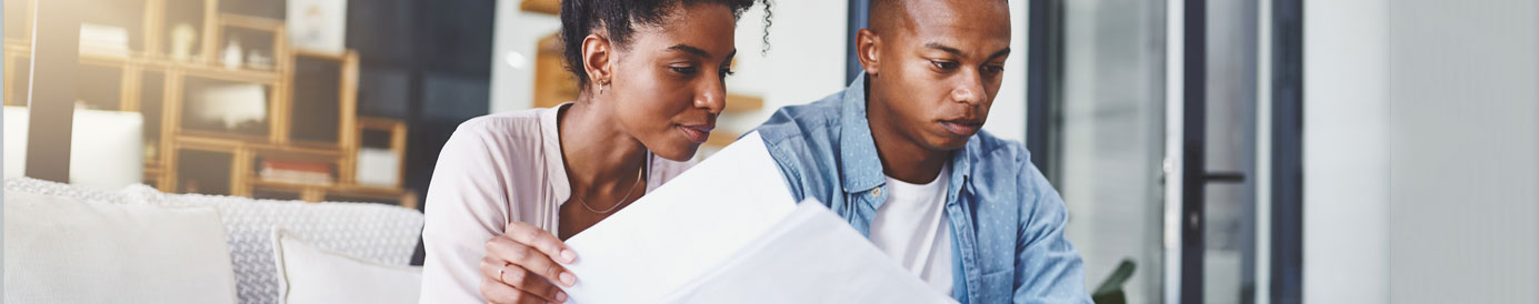 A young couple sits on a couch with documents in hand, looking down with intensity.