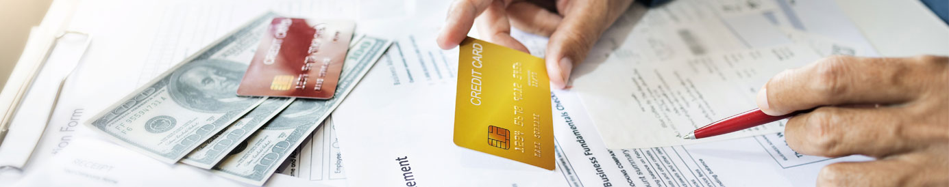 A surface covered in financial statements, a credit card and cash. A person’s arms reach over with a pen and credit card.