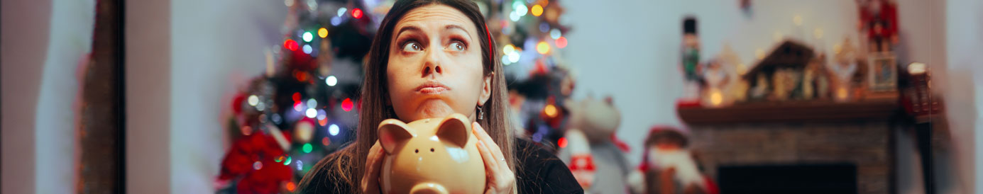 A quizzical looking woman sits in front of a Christmas tree and other holiday decorations with a piggy bank in her hands.