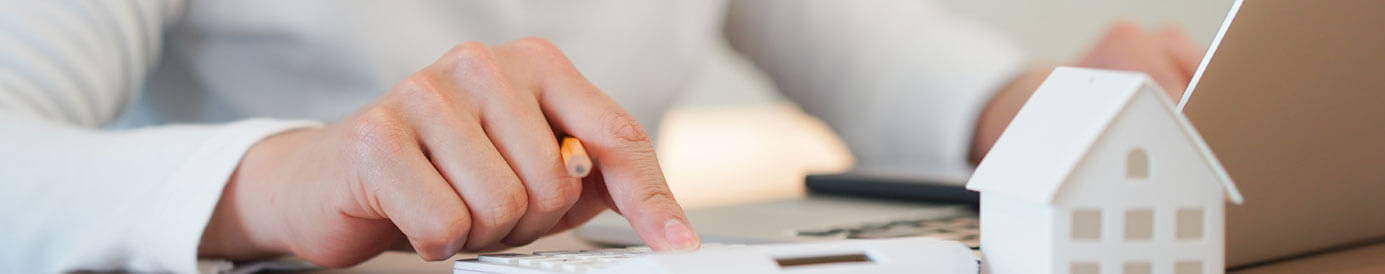 A woman taps on a calculator with her right hand and a computer with her left. A small plastic house in the foreground.