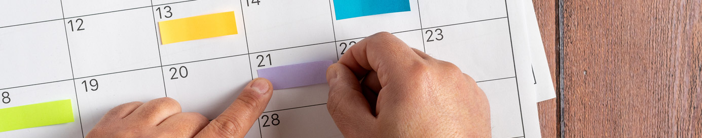 Three months of a calendar are spread across a table. A pair of hands use sticky notes to color-code certain days of the week.