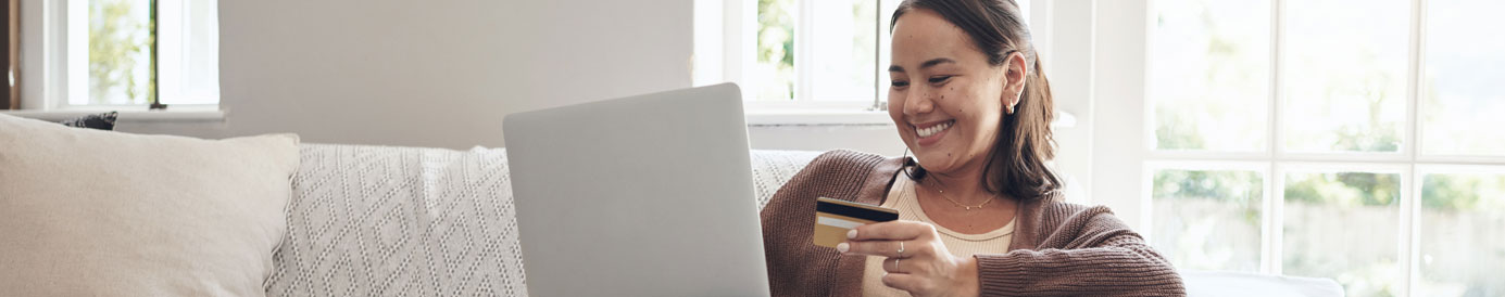 A smiling woman sits on a couch with her laptop on her knee and a credit card in her left hand.