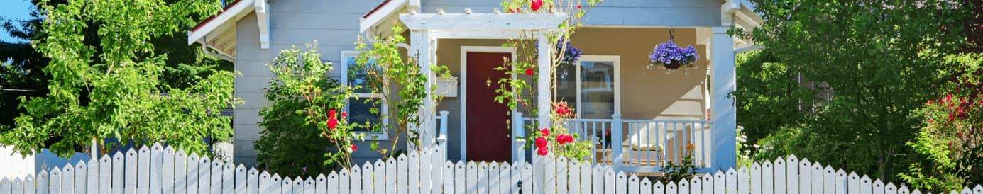 A white picket fence in front of a gray house with a red door