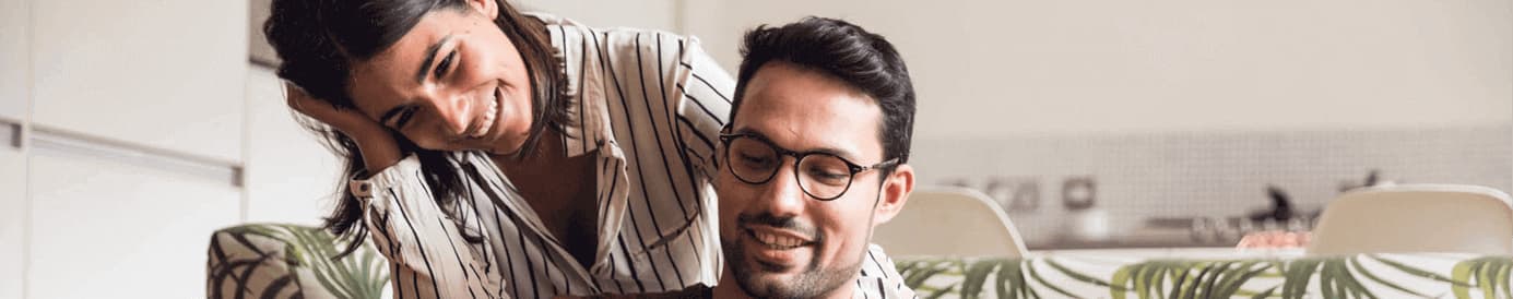 A smiling woman leaning on a man wearing glasses who is smiling while looking and pointing at his laptop