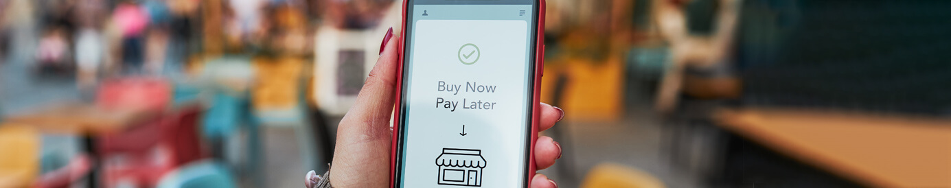 Hands hold a mobile phone. The phone’s screen shows a shop icon with the text 'buy now pay later' and a 'buy now' button