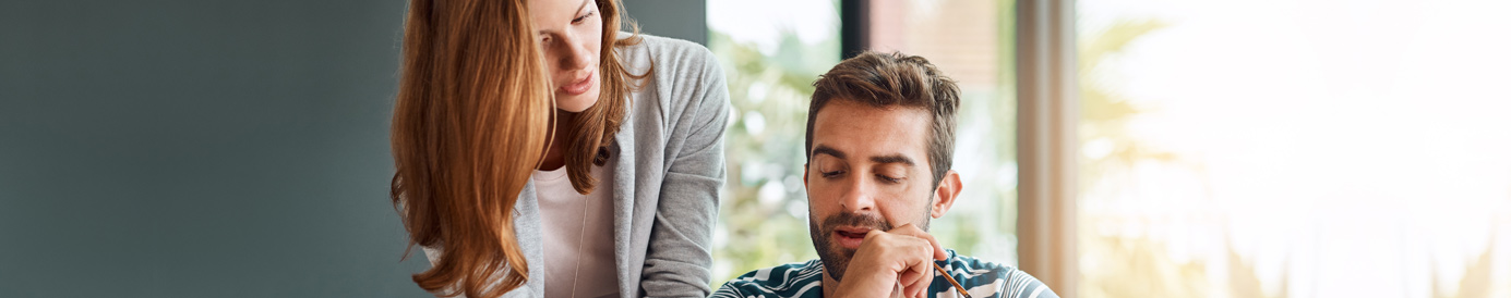 A woman with long red hair stands over a man sitting at a table with a laptop. She points down at a document on the table.