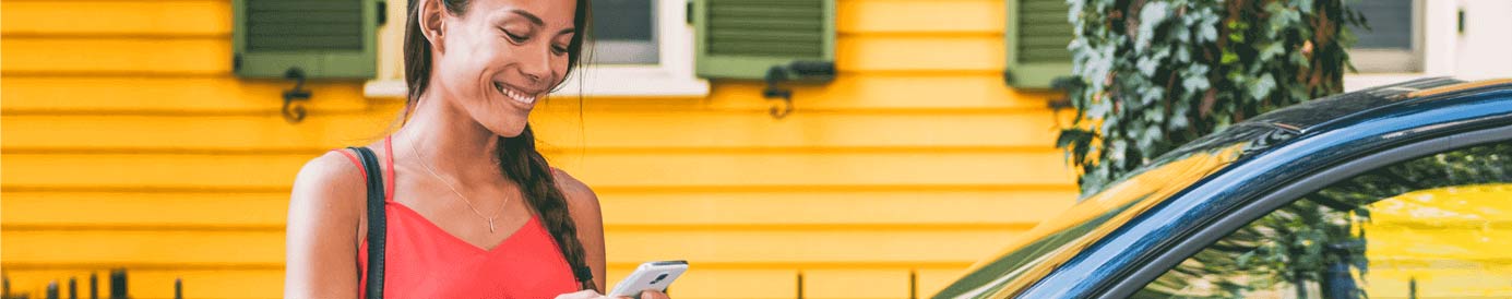 A woman who is standing next to her car and is looking happily at her phone in her hand