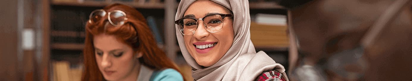 A woman wearing glasses and a hijab smiling and a woman in the background focusing