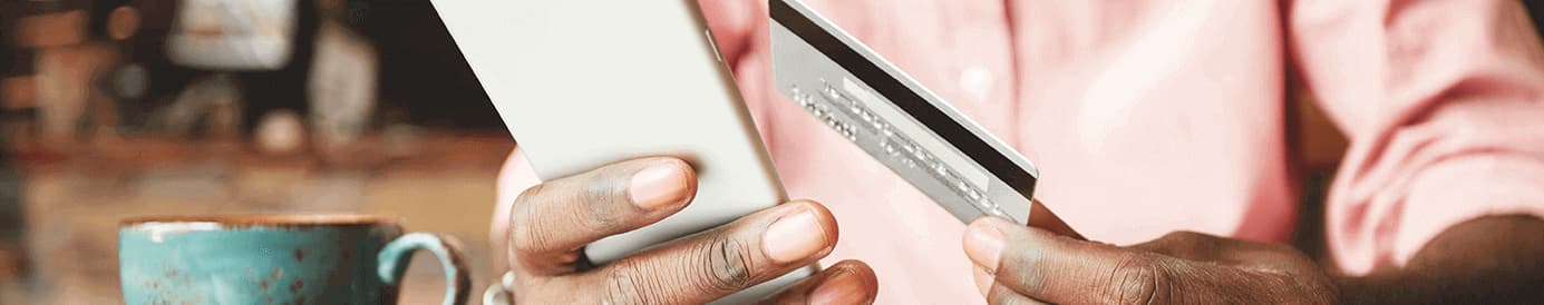A person holds a cellphone in their right hand and a credit card in their left hand as a blue coffee cup sits on the table next to the cellphone