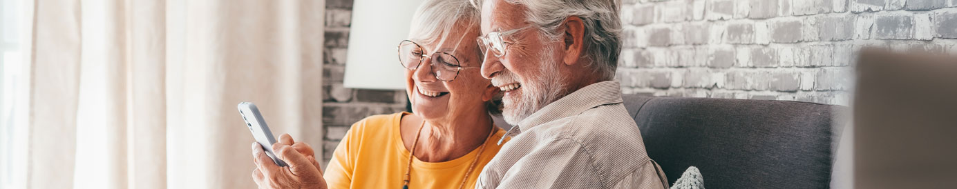 An elderly couple sit on a couch in a brightly lit room with big smiles on their faces while they look at a cell phone.