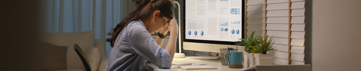 A woman sits at a desk with a computer. She’s distraught, with her elbows on the desk and her head in her hands.