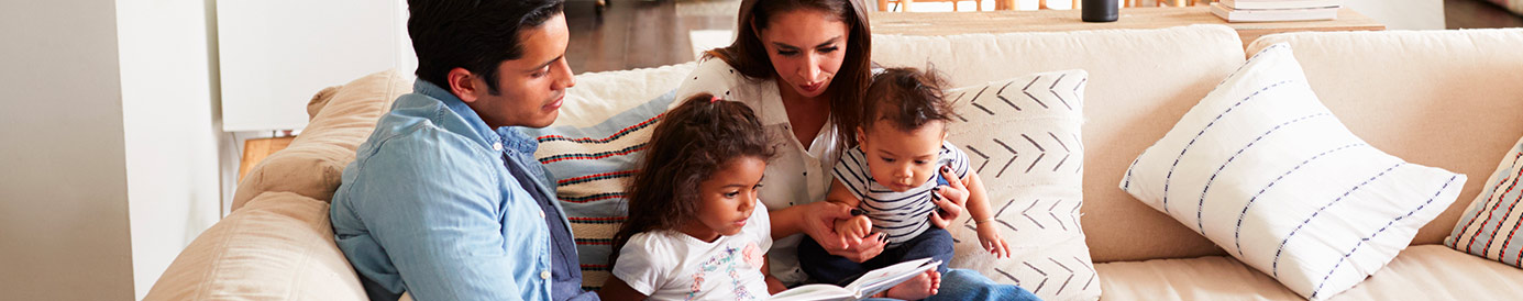 A couple sits in their living room with their two kids. All four of them are sitting close together on a couch and reading a book.