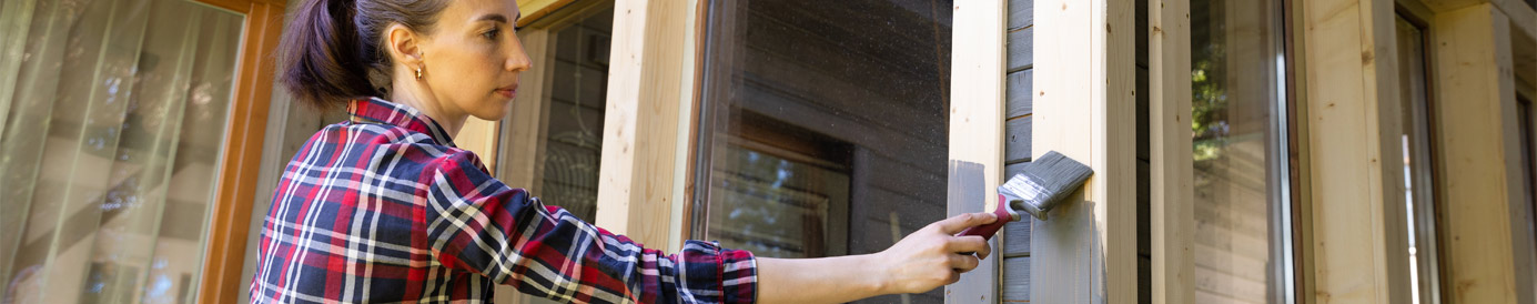 A woman, with a ponytail, stands on a ladder outside a house, reaching out with a paintbrush to paint window trim.