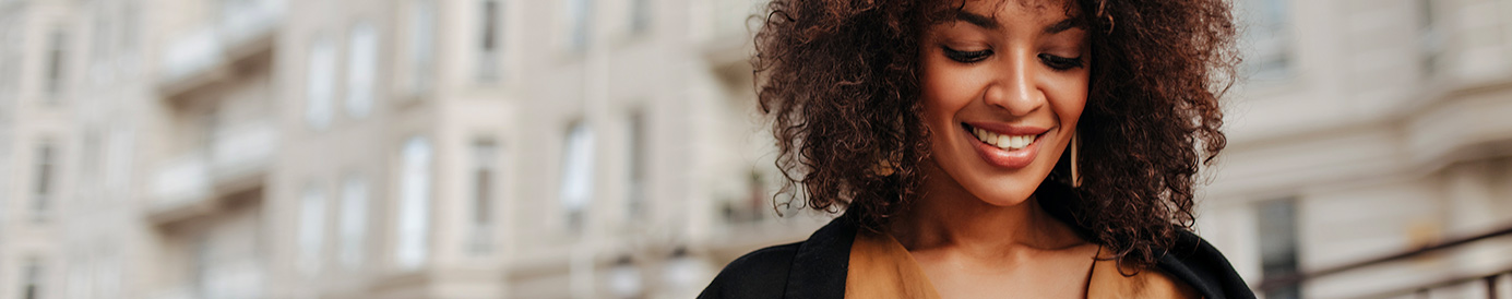 A woman is smiling while looking at her phone. She is standing outside of an apartment complex.