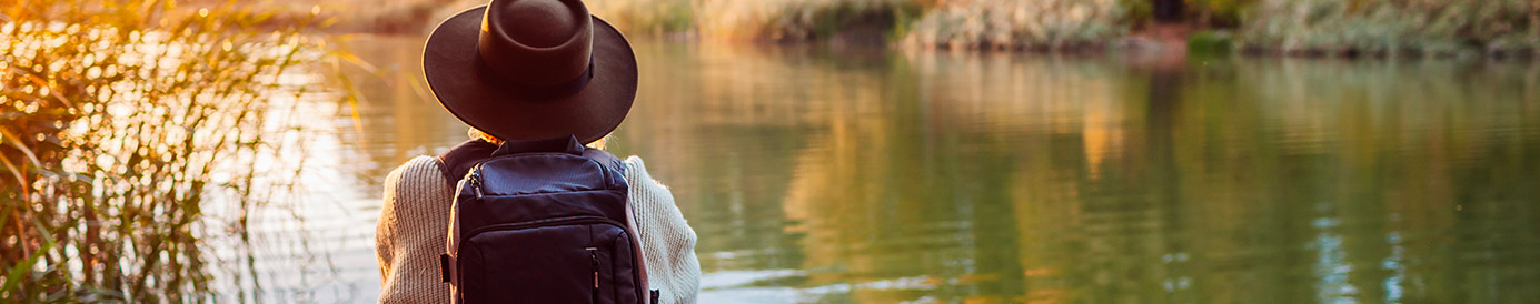 A girl sits on a log right next to a river, looking out at the river. She is wearing a sweater, a flat brim western hat and a backpack.