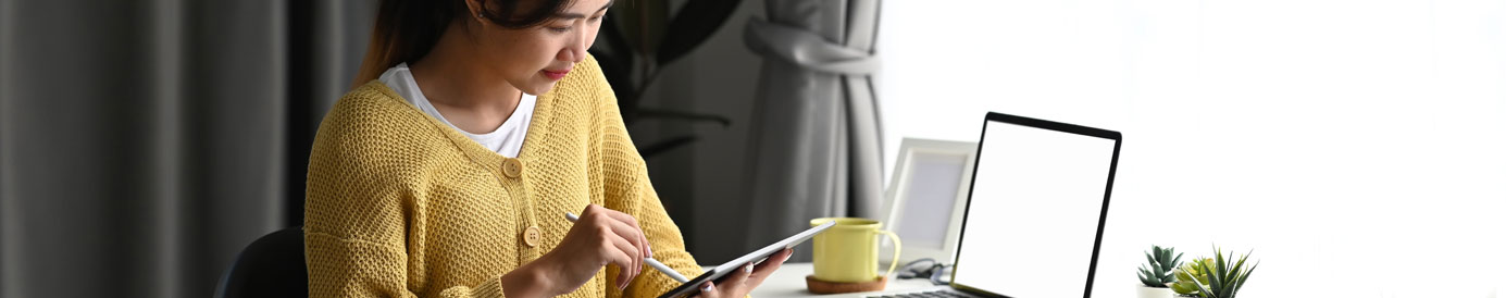 A woman with dark hair and a yellow sweater sits at a desk working on a tablet with a stylus. A laptop rests on the desk.