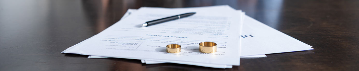 Divorce documents with gold wedding rings and a pen resting on top of them sit on a black surface.