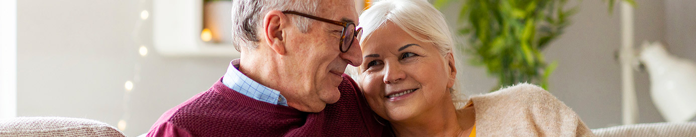 A couple sits together on the couch smiling at each other with white coffee mugs in their hands.