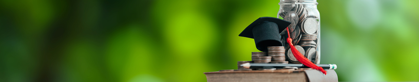 On a wooden surface, a book rests on its side with a jar of change, a pencil, and a small graduation cap sitting on it.