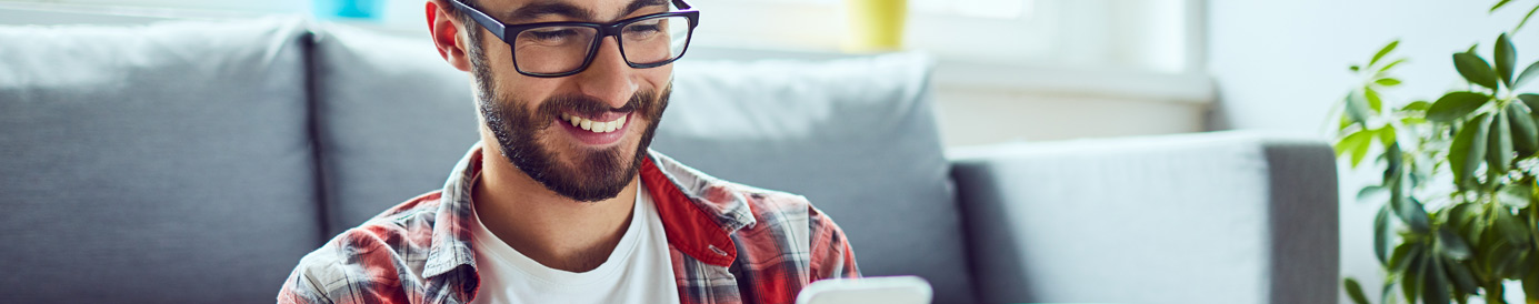 A smiling man with glasses, a beard and short brown hair looks down at his mobile phone as he sits on the floor.