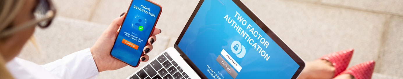 A woman with blonde hair sits with a laptop and mobile phone in hand. Both devices show a multi-factor authentication screen.