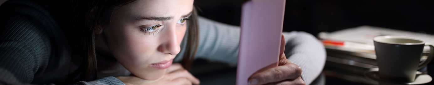 A dark-haired young woman rests her head on a dark surface while looking at a cell phone with a sad look on her face.