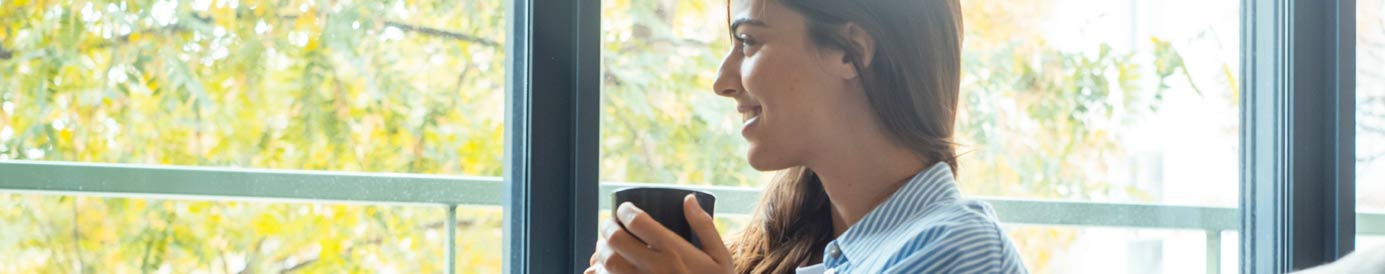 A woman with long brown hair sits smiling with a cup of coffee in her hands as she looks out the window.