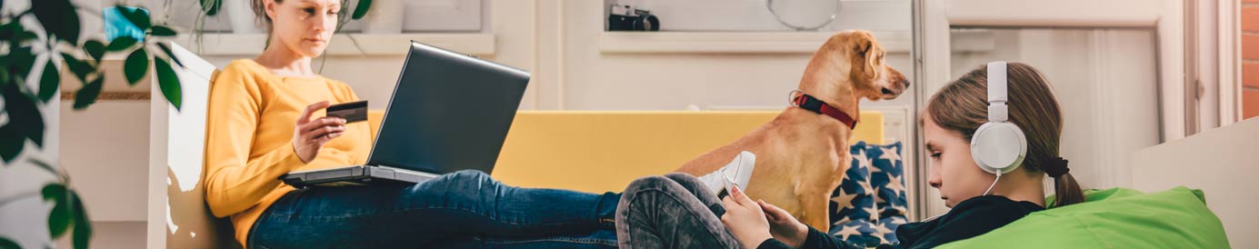 A woman lays on a couch with her laptop and a credit card. A young girl is looking at her phone and a dog sits next to her.