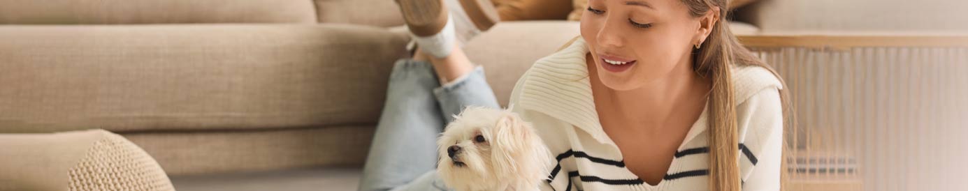 A smiling young woman lays on the floor with her feet up. She looks over at a small white dog that lays next to her.