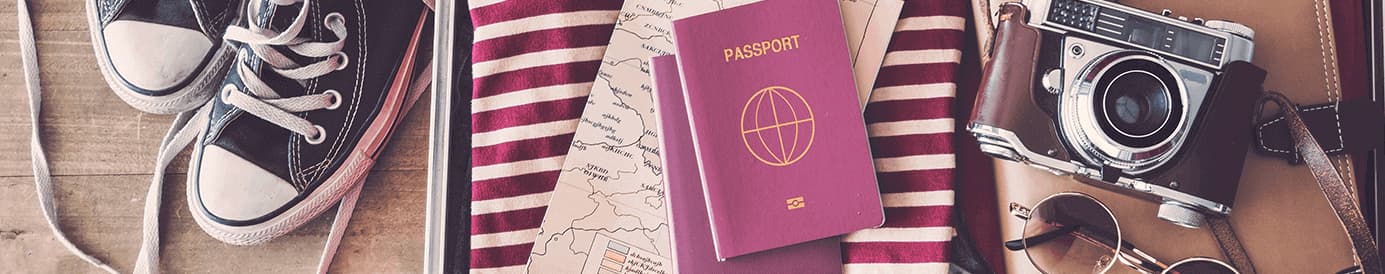 A red passport sitting on top of a map and clothes in a suitcase with a pair of shoes, camera, sunglasses, and a folder off to the side