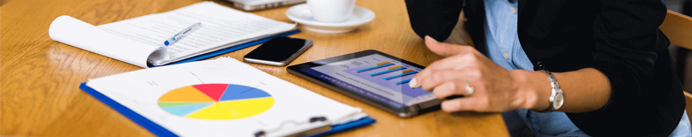 A woman looking down at a graph on a tablet with a pie chart, cellphone, notepad and pen, clsoed laptop and coffee cup also rest on the table