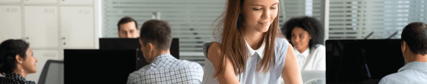 A woman sets up her new desk at work. She puts a small plant on her desk, next to her computer. Several coworkers sit behind her.
