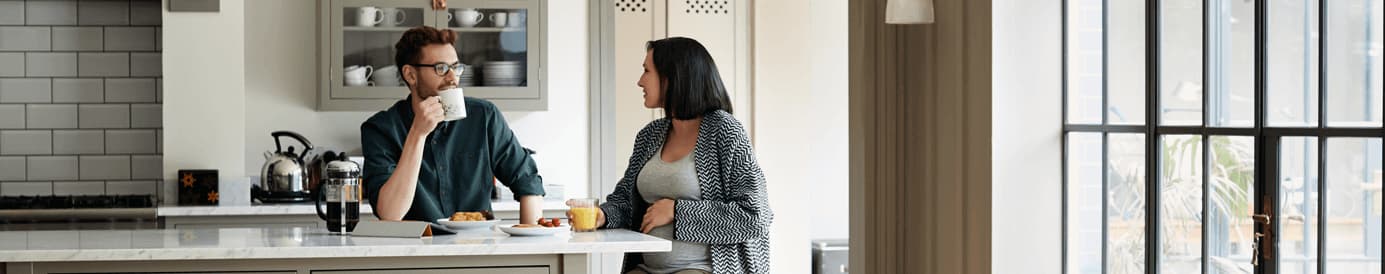 A man wearing glasses and holding a mug and a pregnant woman look at each other while having breakfast