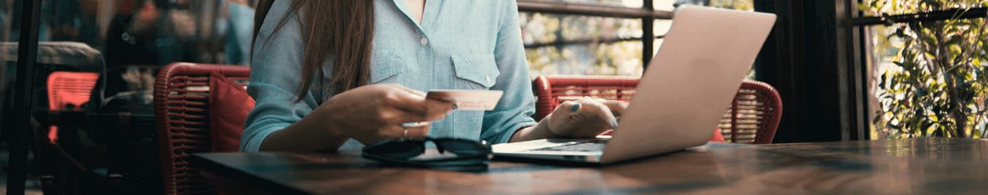 A woman sitting at a table inside a shop is reading her credit card information with a laptop in front of her.