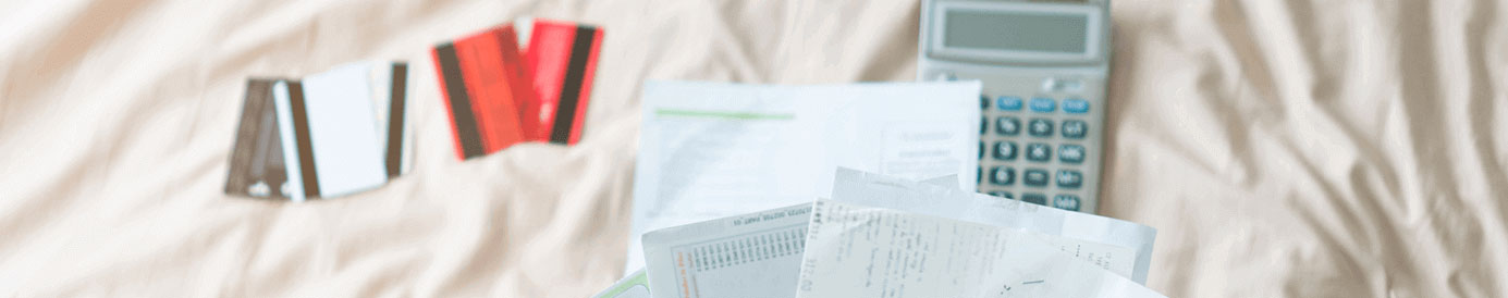 A calculator and 5 credit cards lay spread out on a bed. A person holds up a handful of financial papers above.