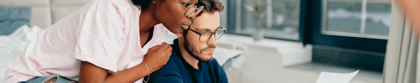 A couple looks over a financial paper together. The man is seated, while the woman leans on him looking over his shoulder.