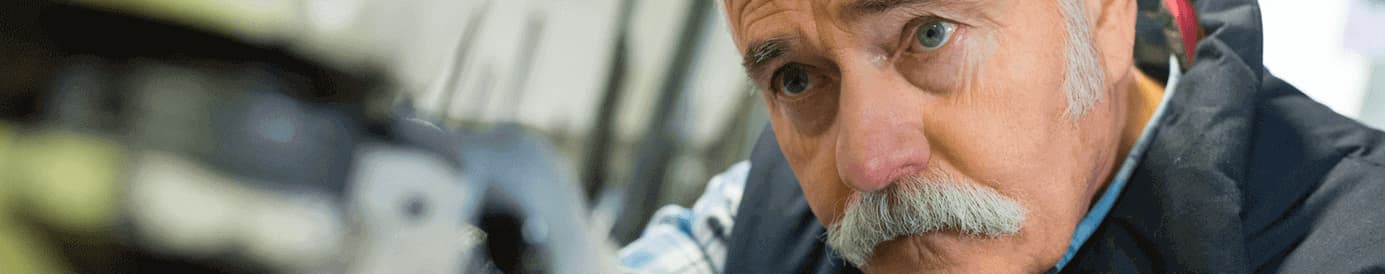 A man in a workshop works on a piece of machinery. He is wearing a striped button down and a work vest.