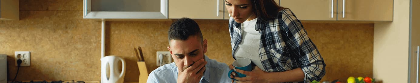 A man sits at a kitchen table looking at a paper and looks distressed. A woman stands over him also looking at the paper.