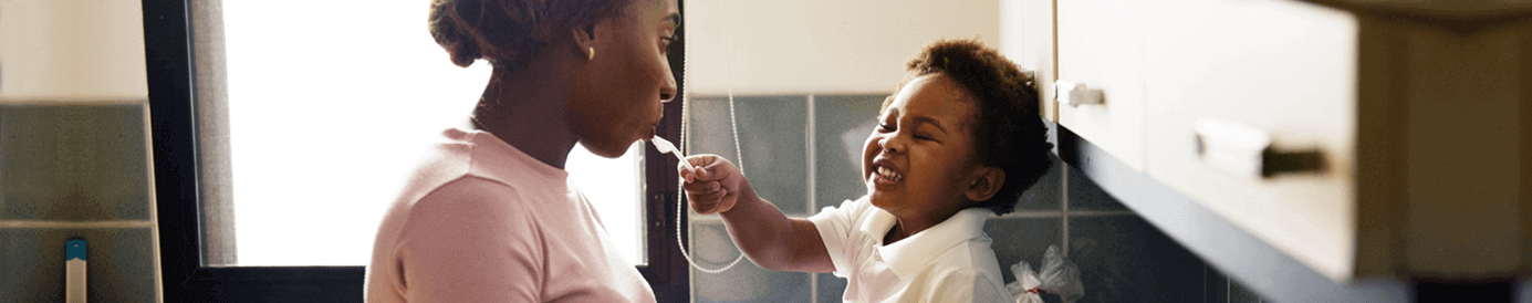 A mom and her child smile at each other in the kitchen. The child sits on top of the counter, while feeding his mom with a spoon.