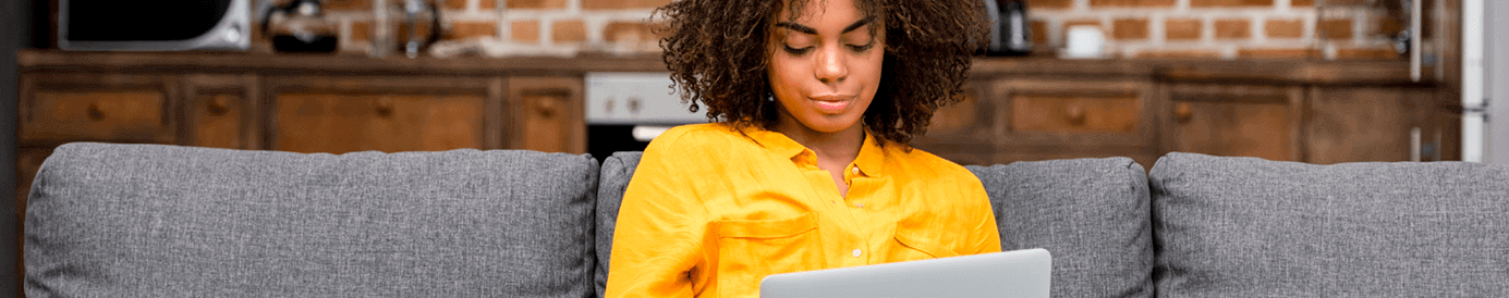 A woman looks at her laptop while sitting on a couch. She is wearing a bright yellow button down.