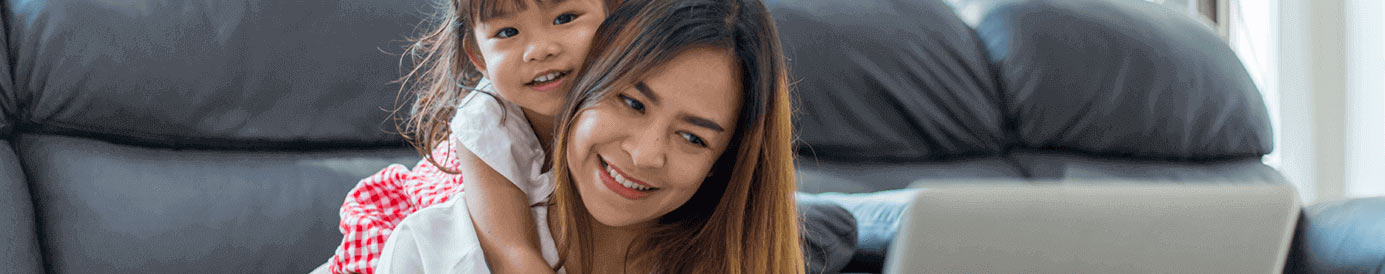 A mom smiling while using her laptop; her daughter is smiling while she hugs her mom.