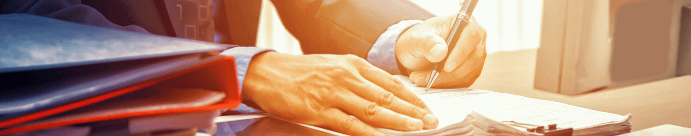 A man signing a pile of papers with a pen in his left hand while a tablet and a pile of binders sit next to the pile of papers on a desk