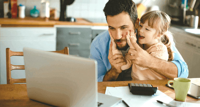 A smiling child holding onto a man's face while he looks at a laptop on a table