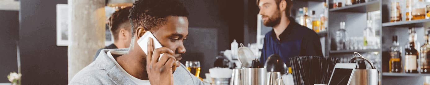 A man sits in a bar with his cellphone to his right ear and his left hand typing on his laptop keyboard