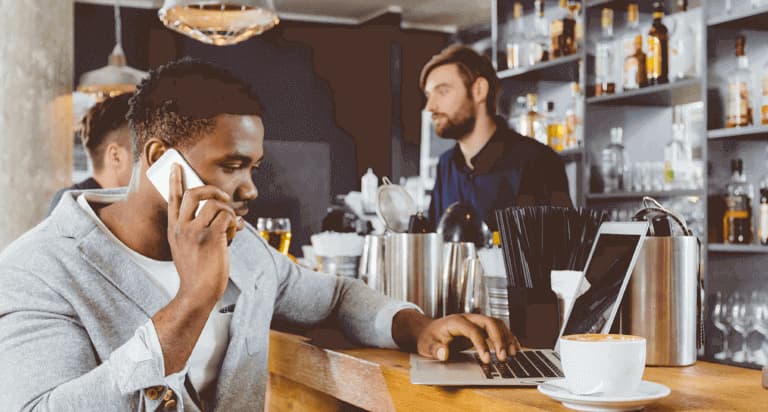 A man sits in a bar with his cellphone to his right ear and his left hand typing on his laptop keyboard