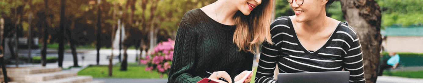 Two women sitting in the grass while the woman on the left holds a book and the woman on the right holds a laptop