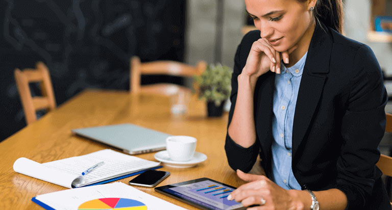 A woman looking down at a graph on a tablet with a pie chart, cellphone, notepad and pen, clsoed laptop and coffee cup also rest on the table